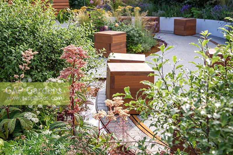 Vue à travers l'espace public, avec des sièges de cube en bois mobiles sur un rail. The Crest Nicholson Livewell Garden, Exposition florale de Hampton Court, 2019.
