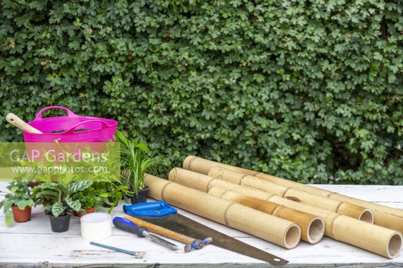 Bambou, scie à main, marteau, ciseau, ruban de masquage, crayon, compost et une variété de petites plantes disposées sur une table