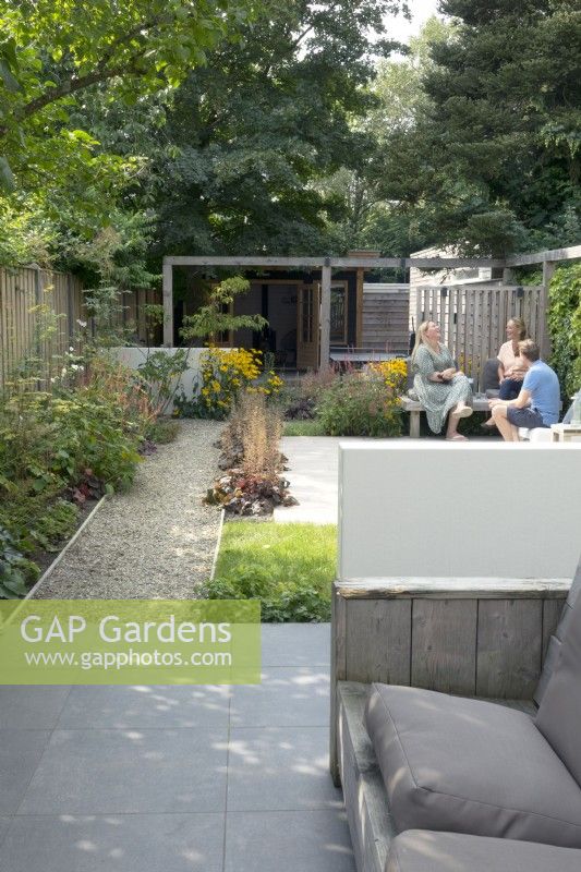 Jardin mitoyen aux murs bas blancs. Vue d'ensemble jardin avec pergola, terrasse, allée, bancs en bois.