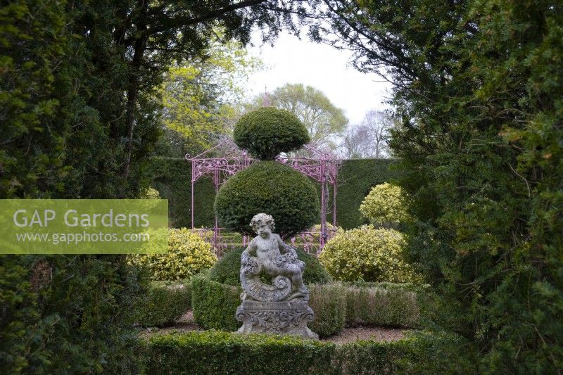 Une statue de chérubin en pierre sculptée baroque entourée d'une haie taillée et d'une topiaire d'if dans le jardin de sculptures à Thenford Gardens and Arboretum, Thenford, Banbury, Oxfordshire, UK
