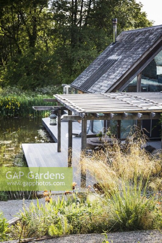Maison conçue par Hugh Wray-McCann à Am Brook Meadow dans le Devon en juin, avec une terrasse en bois le long d'un côté à côté du lac attenant.