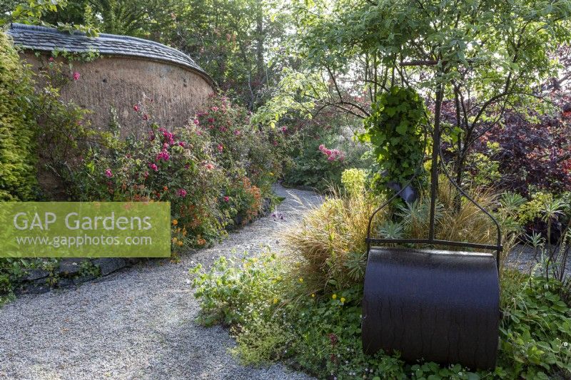 Un chemin de gravier mène à travers un jardin d'été avec un mur en torchis soutenant un parterre de fleurs. Rouleau à gazon en premier plan.