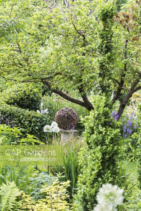 Sphère décorative en métal sur un socle en pierre entourée de plantations luxuriantes dans un jardin de juin