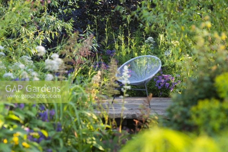 Vue sur les parterres de fleurs jusqu'à la terrasse en bois en bois avec une chaise. RHS Garden pour un avenir vert. Conçu par Jamie Butterworth. RHS Hampton Court Palace Garden Festival Show, juillet 2021
