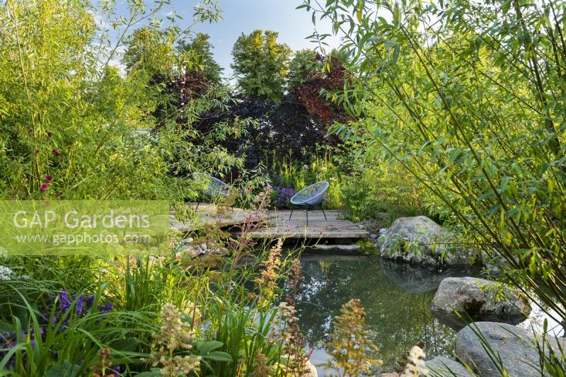 Vue sur l'étang jusqu'au coin salon sur une terrasse en bois en bois. RHS Garden pour un avenir vert. Conçu par Jamie Butterworth. RHS Hampton Court Palace Garden Festival Show, juillet 2021