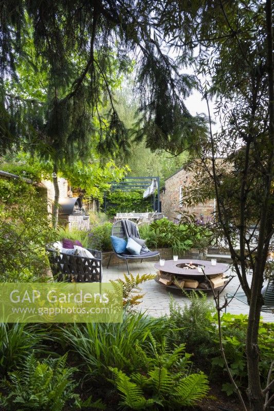 Vue sur jardin boisé avec plantes tolérantes à l'ombre et coin salon en contrebas avec chaises autoportantes et foyer.