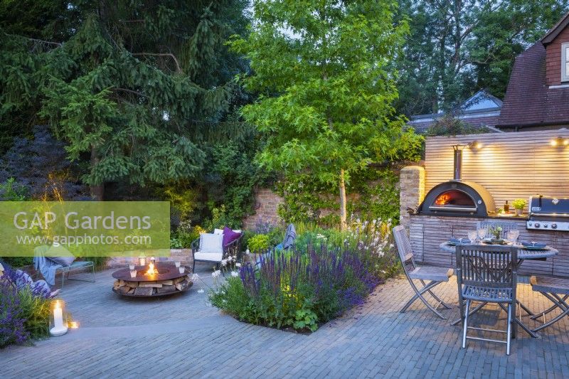 Jardin en contrebas illuminé et coin repas extérieur avec table en bois, chaises et unité de cuisine sur mesure avec four à pizza intégré et barbecue la nuit.