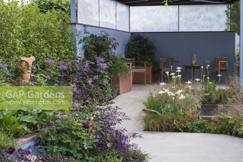 Un sanctuaire serein avec un coin salon couvert à côté d'un étang planté d'Iris 'White Swirl', et entouré de plantations herbacées dans les tons de vert, violet, bronze et bleu.