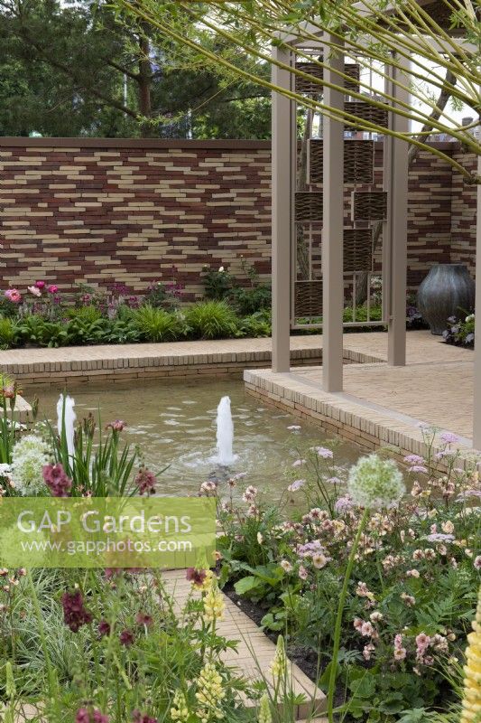 Un bassin en contrebas avec jets d'eau est bordé de parterres herbacés plantés d'une tapisserie de lupins, alliums, geums, pimpinella, verbascums et graminées ornementales.