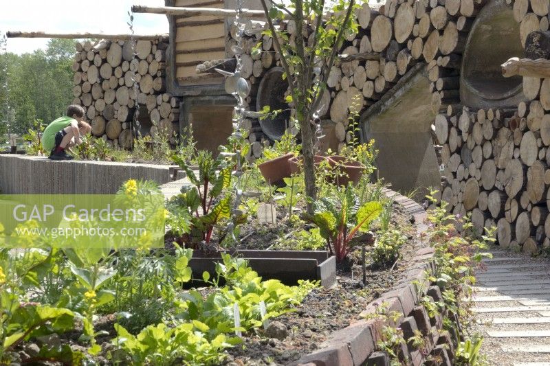 Légumes plantés dans des pots de pierres recyclées et drainage fait de vieilles cuillères en fer. Mur de troncs d'arbres empilés et de tuyaux de drainage en béton.