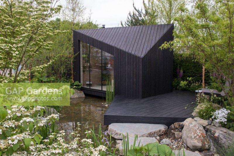 Une cabane en mélèze carbonisé avec terrasse en bois surplombe un étang entouré de Betula nigra - A Garden Sanctuary by Hamptons, RHS Chelsea Flower Show 2022