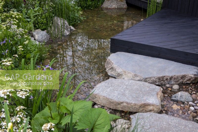 De gros rochers de pierre mènent à une terrasse en bois carbonisé donnant sur un étang avec Iris et Viburnum - A Garden Sanctuary by Hamptons, RHS Chelsea Flower Show 2022