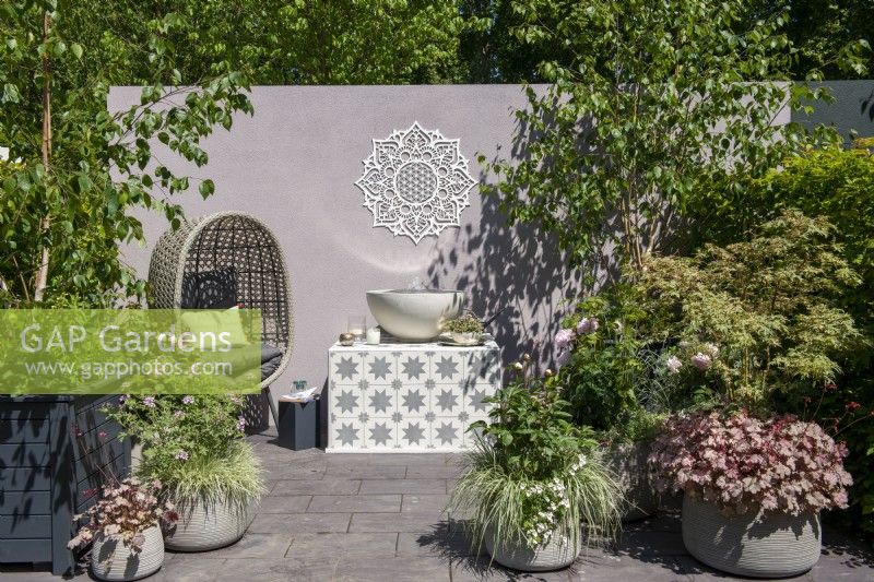 Chaise en rotin à côté d'une pièce d'eau sur un support carrelé entouré de plantes en pots - The Mandala Garden, RHS Chelsea Flower Show 2022 - Médaille d'argent