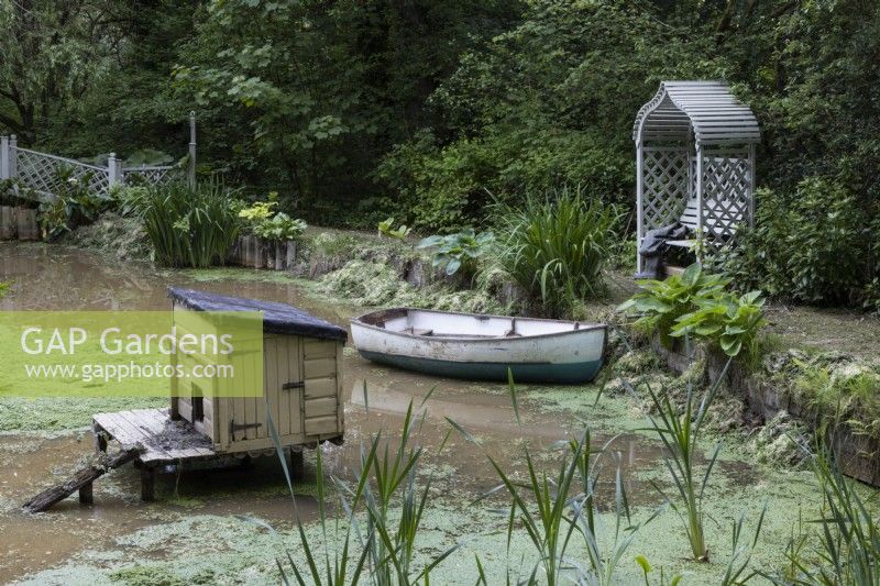 Une maison de canard en bois se trouve sur un étang à côté d'un bateau à rames en bois avec un siège de tonnelle en bois à l'arrière-plan. Lewis Cottage, jardin NGS Devon. Le printemps.