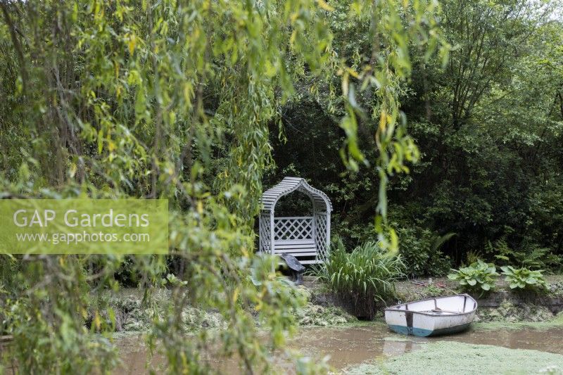 Un bateau à rames en bois sur un étang avec un siège de tonnelle en bois sur le bord et une paire d'échassiers à côté du siège avec un feuillage de saule en premier plan. Lewis Cottage, jardin NGS Devon. Le printemps.