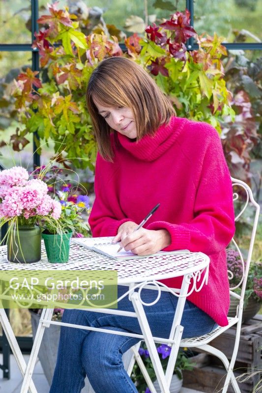 Femme assise à une table écrivant dans une serre qui a été remplie de diverses plantes et pots mélangés