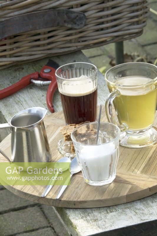 Café, thé, sucre et lait sur plateau en bois et sécateurs sur banc en bois.