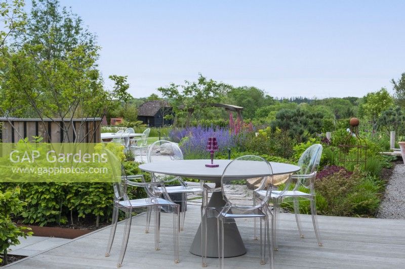 Une terrasse en bois surélevée surplombe des plates-bandes de vivaces et de petits arbres.