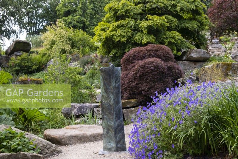 Une sculpture en métal entourée de géranium, d'arbres et de formations rocheuses à Paxton's Rock Garden, Chatsworth House and Garden.