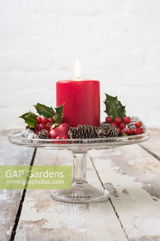 Décoration de table de Noël avec bougie rouge, cônes et feuillage Ilex et baies sur présentoir à gâteaux en verre vintage
