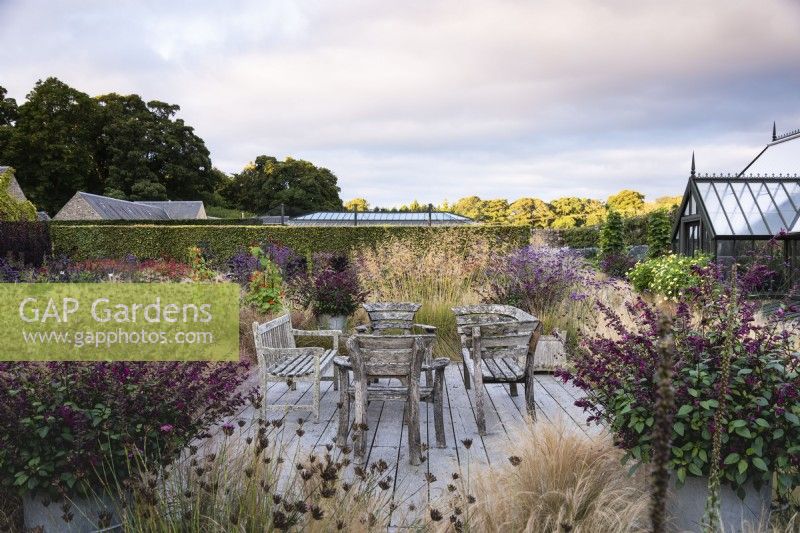 Espace aménagé avec des sièges en bois entourés de graminées ornementales et de plantes vivaces à floraison tardive, dont Verbena bonariensis et des pots de salvias.