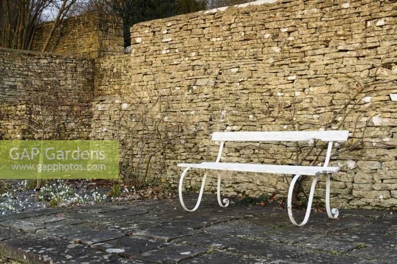 Un banc blanc soutenu par un mur de pierre de Cotswold à Cotswold Farm Gardens en février.