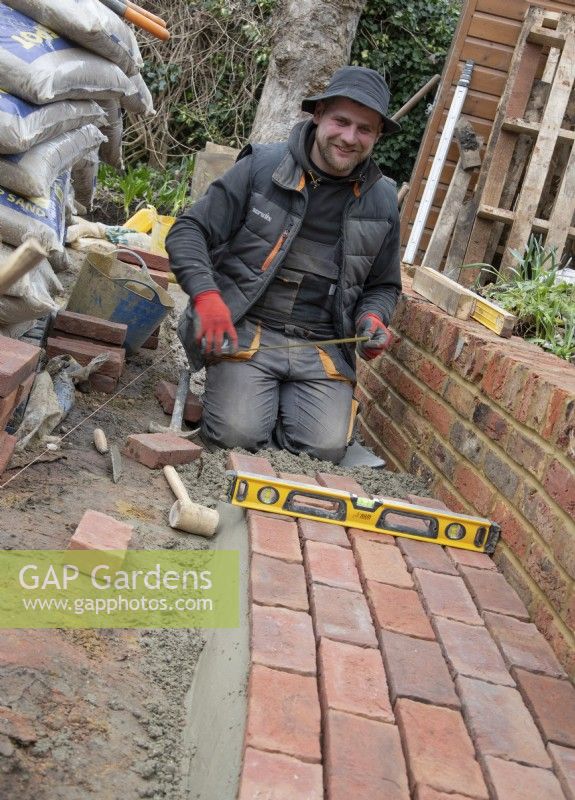 Un ouvrier posant des briques pour un chemin de pierre incurvé autour d'un parterre de fleurs surélevé lors de la rénovation d'un petit jardin de Londres.