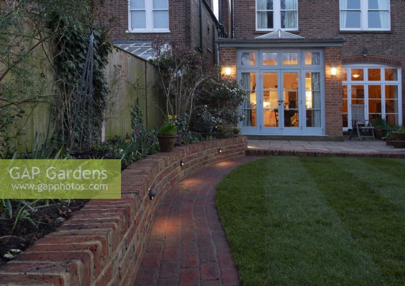 Un jardin londonien après une cure de jouvence avec des éclairages vers le bas sur un mur de briques, un chemin de briques autour d'un parterre de fleurs surélevé et du gazon nouvellement posé.