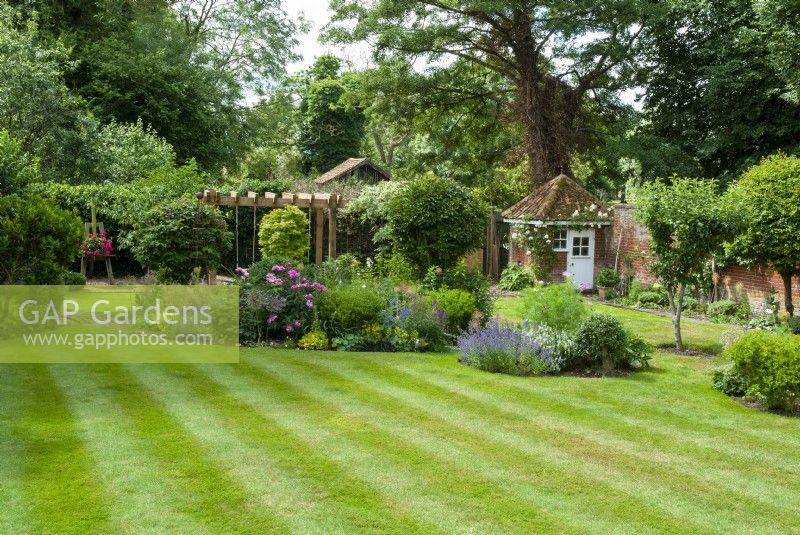 Vue sur grande pelouse à rayures de parterres de fleurs, arbustes, pergola et bâtiment de jardin au-delà - Journée des jardins ouverts, Easton, Suffolk