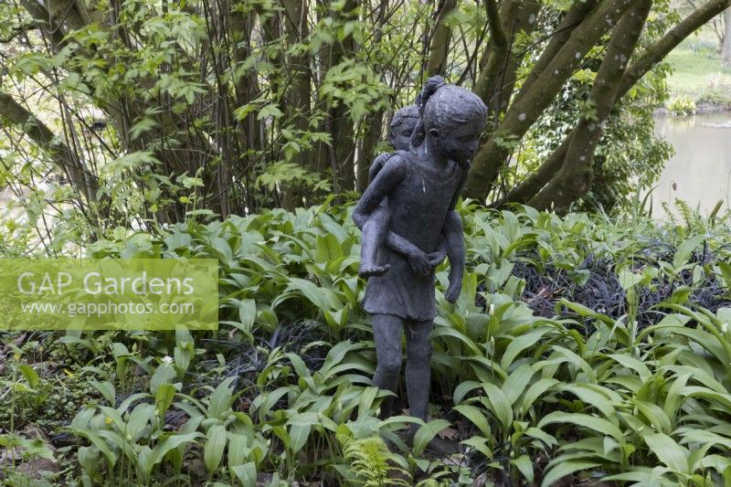 Une statue d'une jeune fille portant un petit enfant, sur le dos parmi la croissance printanière de l'ail sauvage. Jardins de Marwood Hill, Devon. Printemps. Dévon.