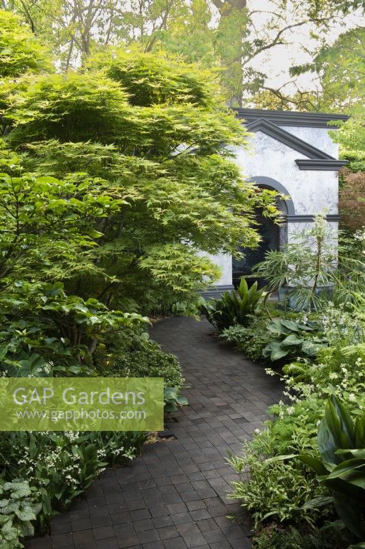 Une allée en blocs de chêne bordée d'érables et de vivaces feuillues mène à un bâtiment de jardin.