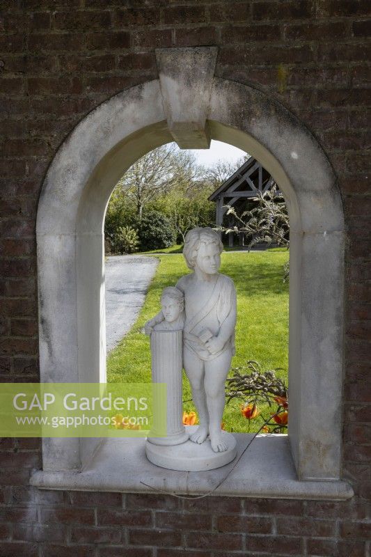 Statue en pierre d'un personnage debout avec un bras autour d'un buste sur un piédestal, installée dans une arche dans un mur de briques avec vue sur des pelouses et un bâtiment à pans de bois. Trago Mills montrent les jardins, Devon, Royaume-Uni. Peut. Printemps