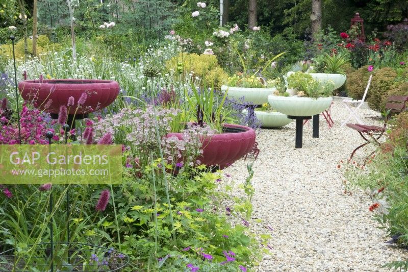 Vasques décoratives roses et vertes sur un socle planté de diverses plantes réparties sur le jardin avec des galets.