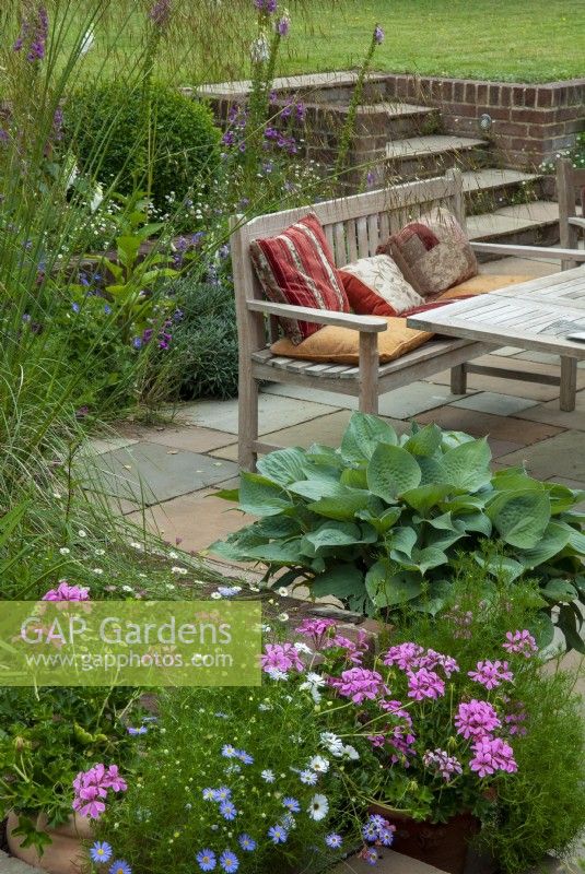 Patio en contrebas avec table, siège et coussins et plates-bandes surélevées de plantes vivaces. Étapes menant à la pelouse à un niveau supérieur - Open Gardens Day, Tuddenham, Suffolk