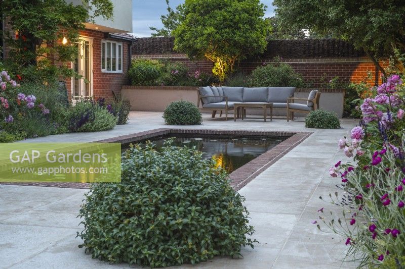 Eclairage de jardin sur terrasse avec bassin rectangulaire et mobilier, boules topiaires Pittosporum pour dissimulation de la lumière et parterres de Roses