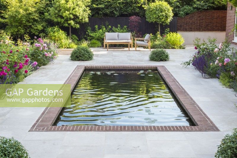 Piscine rectangulaire bordée de briques donnant sur terrasse dallée de grès avec salon de jardin et parterres de rosiers.