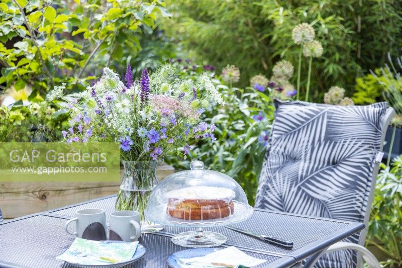 Table de jardin avec bouquet de fleurs, boissons chaudes, assiettes et gâteau fraîchement sorti du four