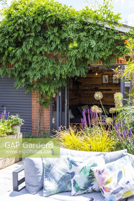 Maison d'été au bout du jardin avec des glycines poussant autour de l'avant derrière un canapé et un parterre de fleurs surélevé