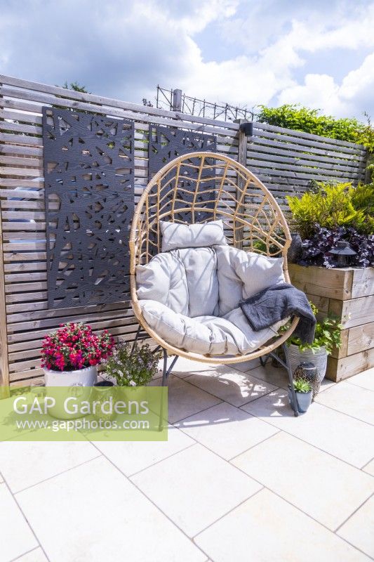 Chaise oeuf en osier sur patio carrelé blanc avec panneaux décoratifs sur clôture et Fuchsia, Thym, Alchemilla et sempervivum en pot au sol