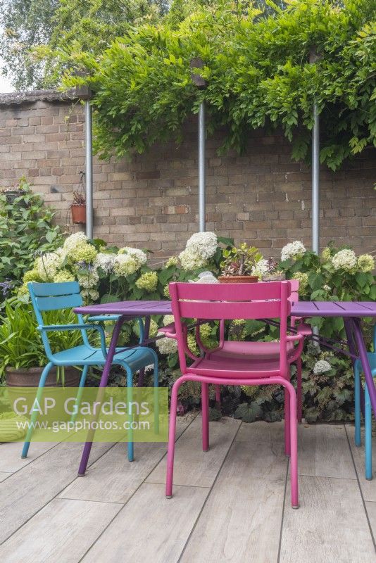 Table et chaises colorées sur un petit jardin urbain clos et pavé avec supports de poteaux d'échafaudage avec glycine et hortensia 'Annabelle'