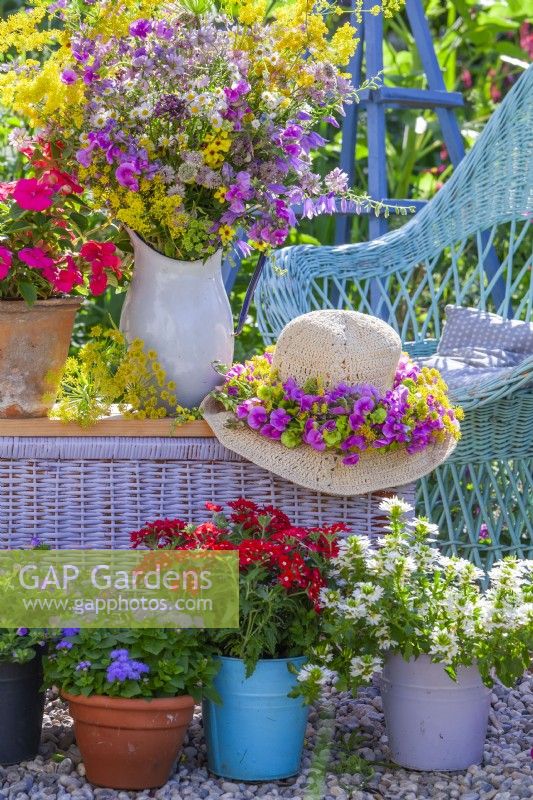 Présentation de pots avec fleurs de balcon, chapeau avec couronne et bouquet de fleurs de pois de senteur et de fleurs sauvages dans un pichet en émail.