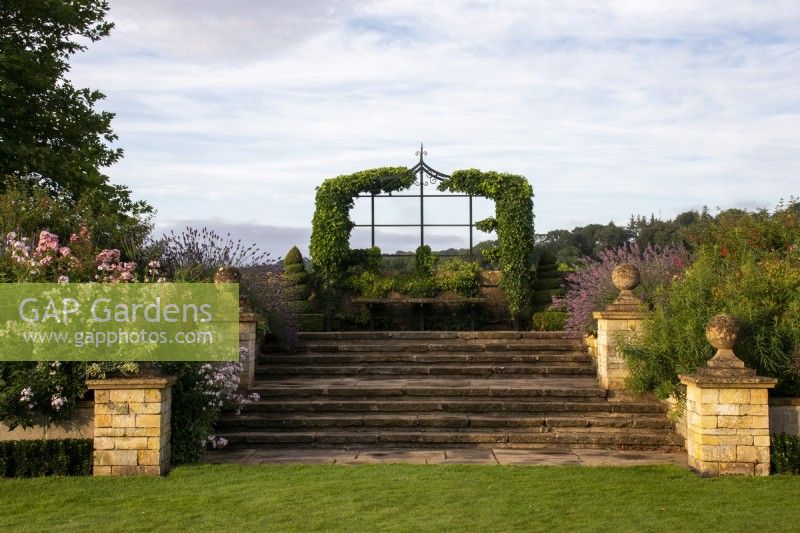 De larges marches et des piliers en pierre surmontés de fleurons ronds mènent à un siège sous une tonnelle recouverte de lierre sur la promenade surélevée du Bourton House Garden, Gloucestershire.