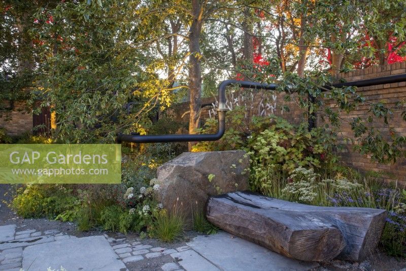 Jardin urbain éclairé la nuit avec des plantations mixtes de plantes vivaces : un coin salon avec banc en tronc d'arbre sculpté, un mur de briques et une sculpture de tuyaux recyclés et récupérés traverse le jardin