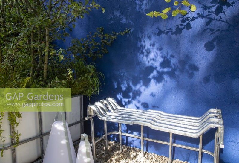 Banquette en métal contemporaine moderne et recyclée, fabriquée à partir d'un IBC - cadre de pot en vrac intermédiaire - sur un patio à surface en gravier - mur enduit peint en bleu