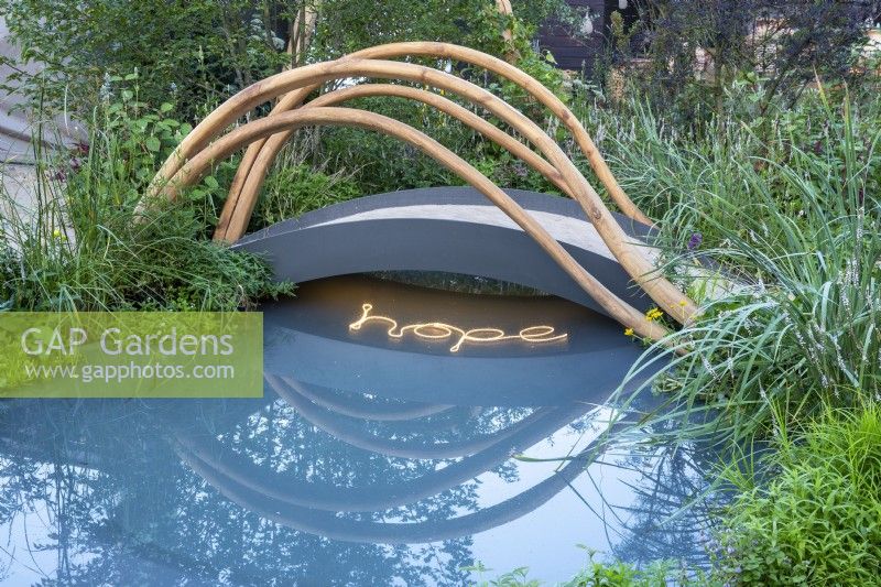 Un petit pont en arc métallique contemporain et moderne avec des rails en bois sculpturaux au-dessus d'un étang avec l'ESPOIR écrit en éclairage dans l'eau - marges d'eau