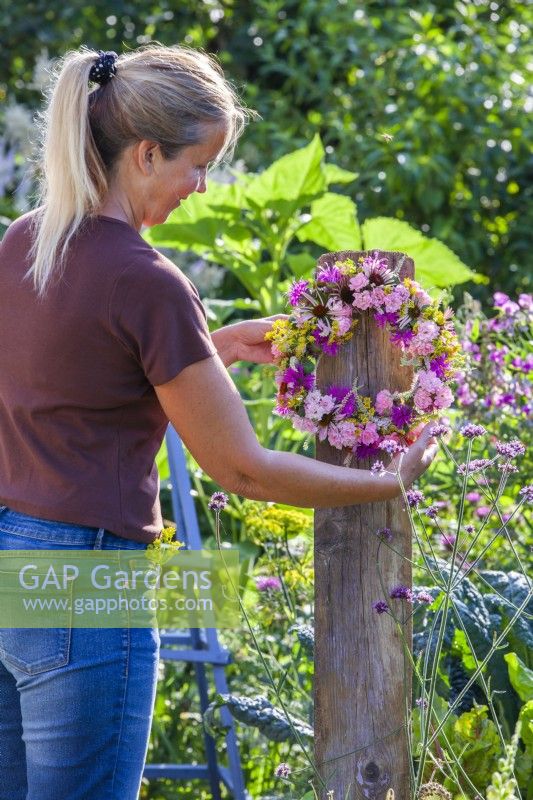 Une femme accroche une couronne principalement rose composée d'échinacée, de rose, de fenouil, de monarda et de veronicastrum sur un support en bois 