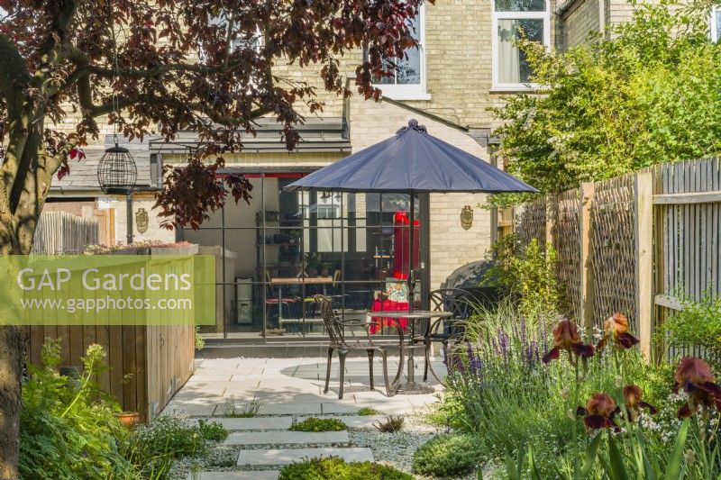 Vue sur terrasse dans un petit jardin de ville contemporain avec parasol sur une table et des chaises et un magasin de jardinage en bois. Juin 