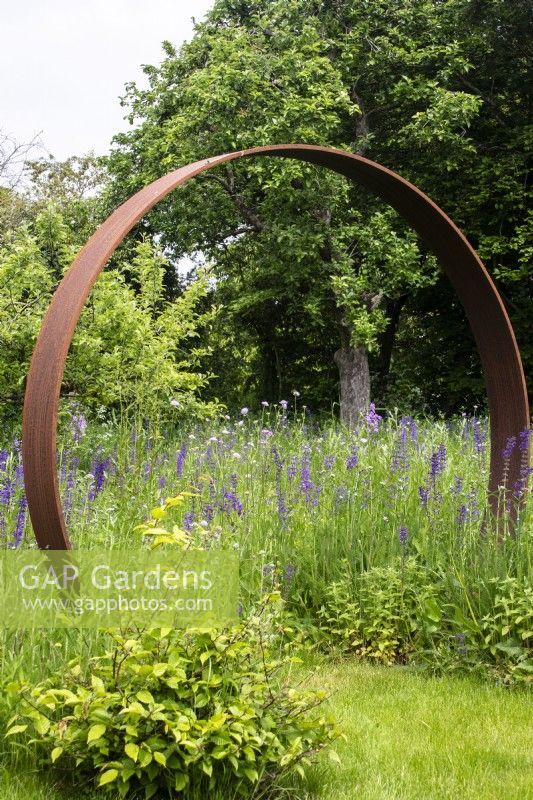 Une porte lunaire en acier Corten marque l’entrée d’une prairie de fleurs sauvages vivaces. 