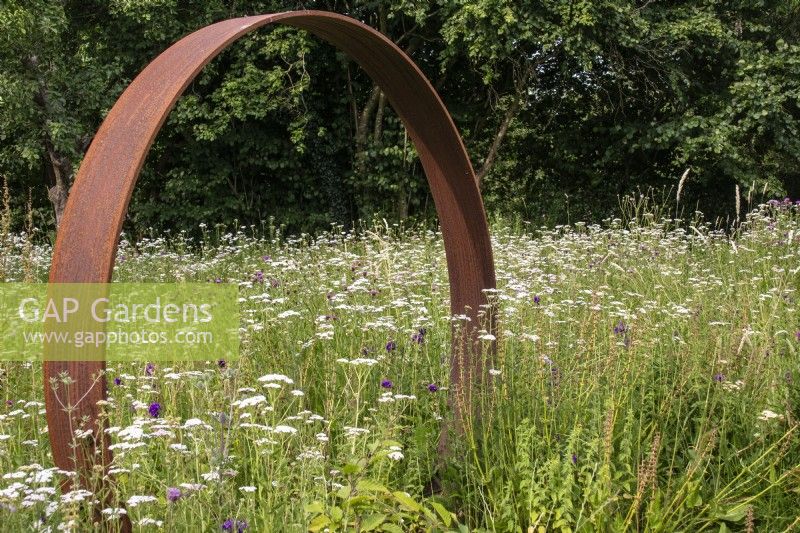 Une porte lunaire en acier Corten se dresse dans une prairie vivace avec Achillea millefolium. 