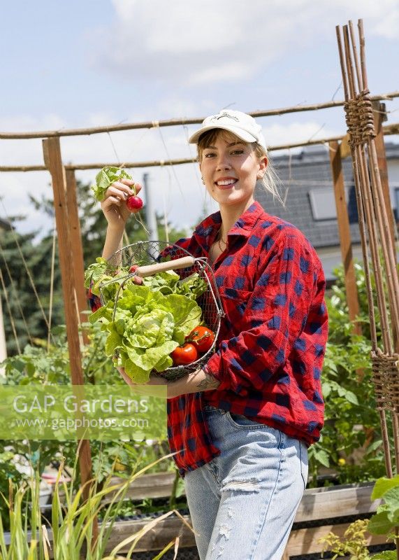 Jeune femme tenant un treillis métallique de radis, de laitue et de tomates dans un potager urbain, été août 